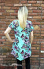 Woman modeling back view of the Hope Pattern tunic length, v hem is about mid thigh level, fabic is blue with florals and butterflies.